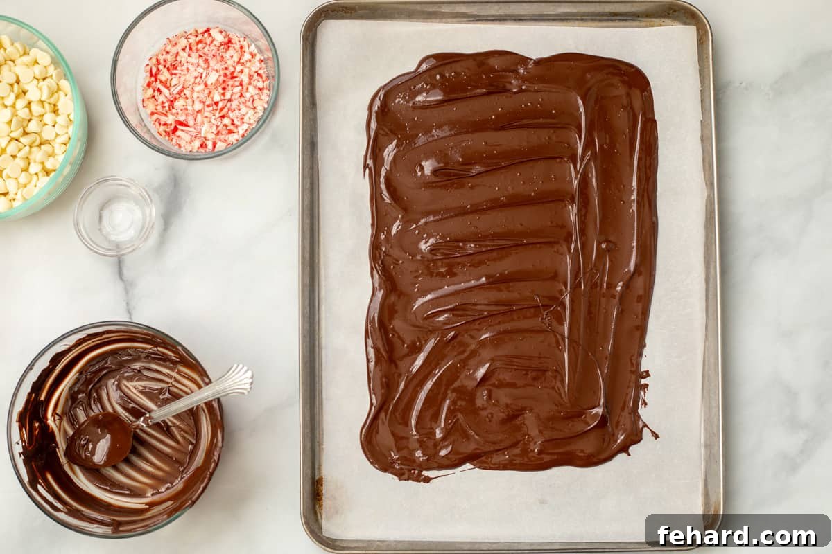 Melted dark chocolate evenly spread out on a parchment-lined sheet pan in a 9x13 inch rectangular shape, forming the base of the peppermint bark.