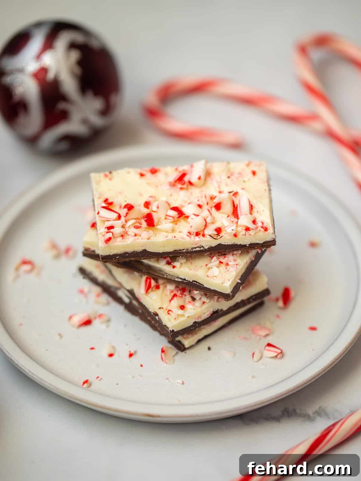 Beautifully stacked pieces of homemade Peppermint Bark on a plate, with candy canes and Christmas decorations in the background, showcasing the festive treat.