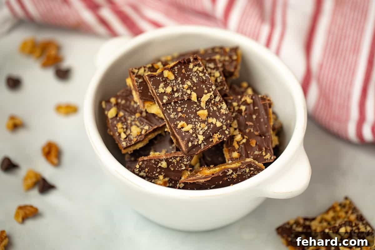 A bowl brimming with broken pieces of Saltine Cracker Toffee, with more walnuts and chocolate chips visible on the countertop.