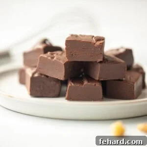 Fudge cut into cubes on white plate with nuts and chocolate chips in background.