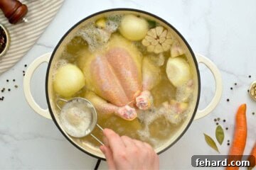 Spoon removing scum from simmering chicken soup in large soup pan.