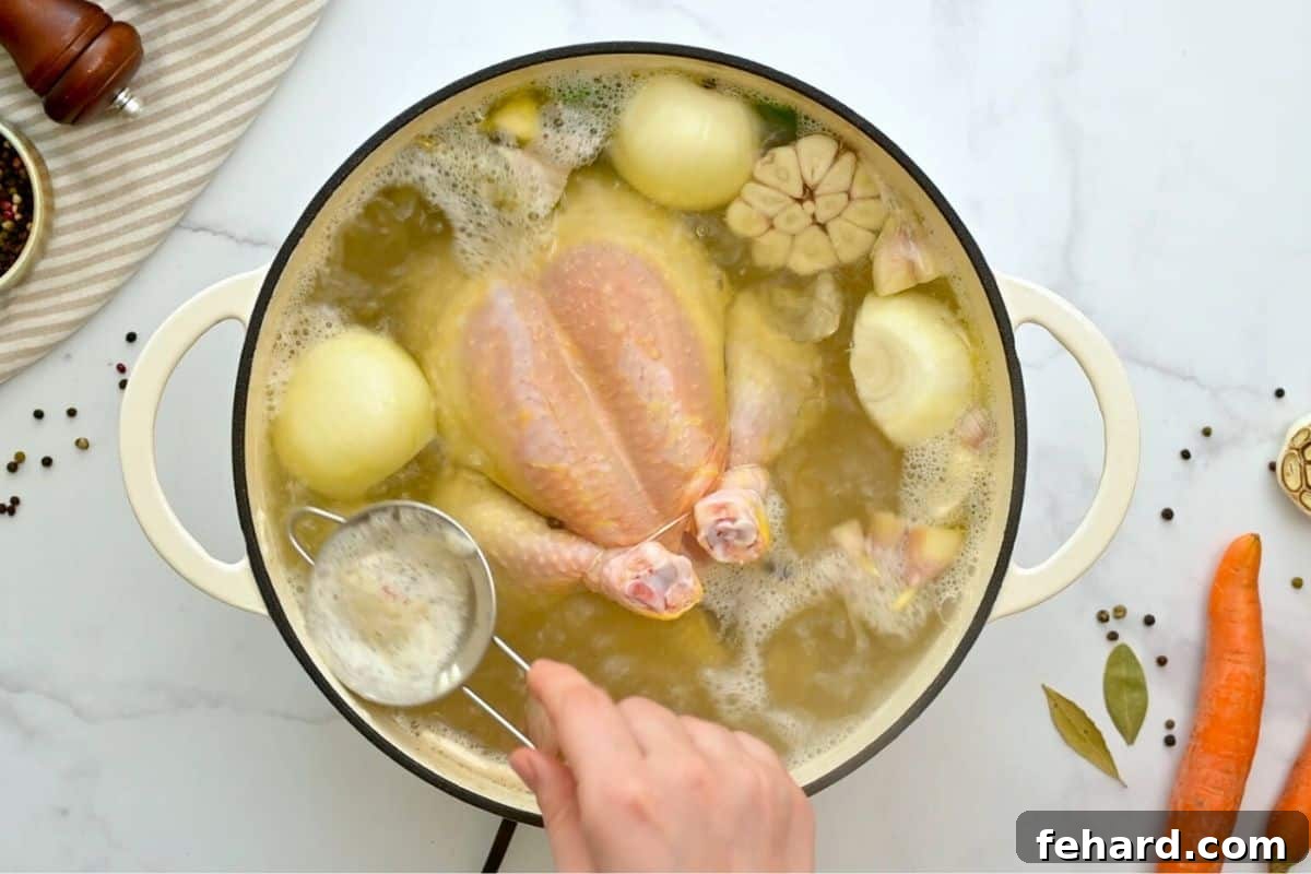 Spoon removing scum from simmering chicken soup in large soup pan.