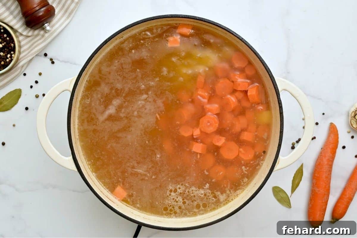 Simmering chicken broth with carrots and potatoes inside large soup pan.