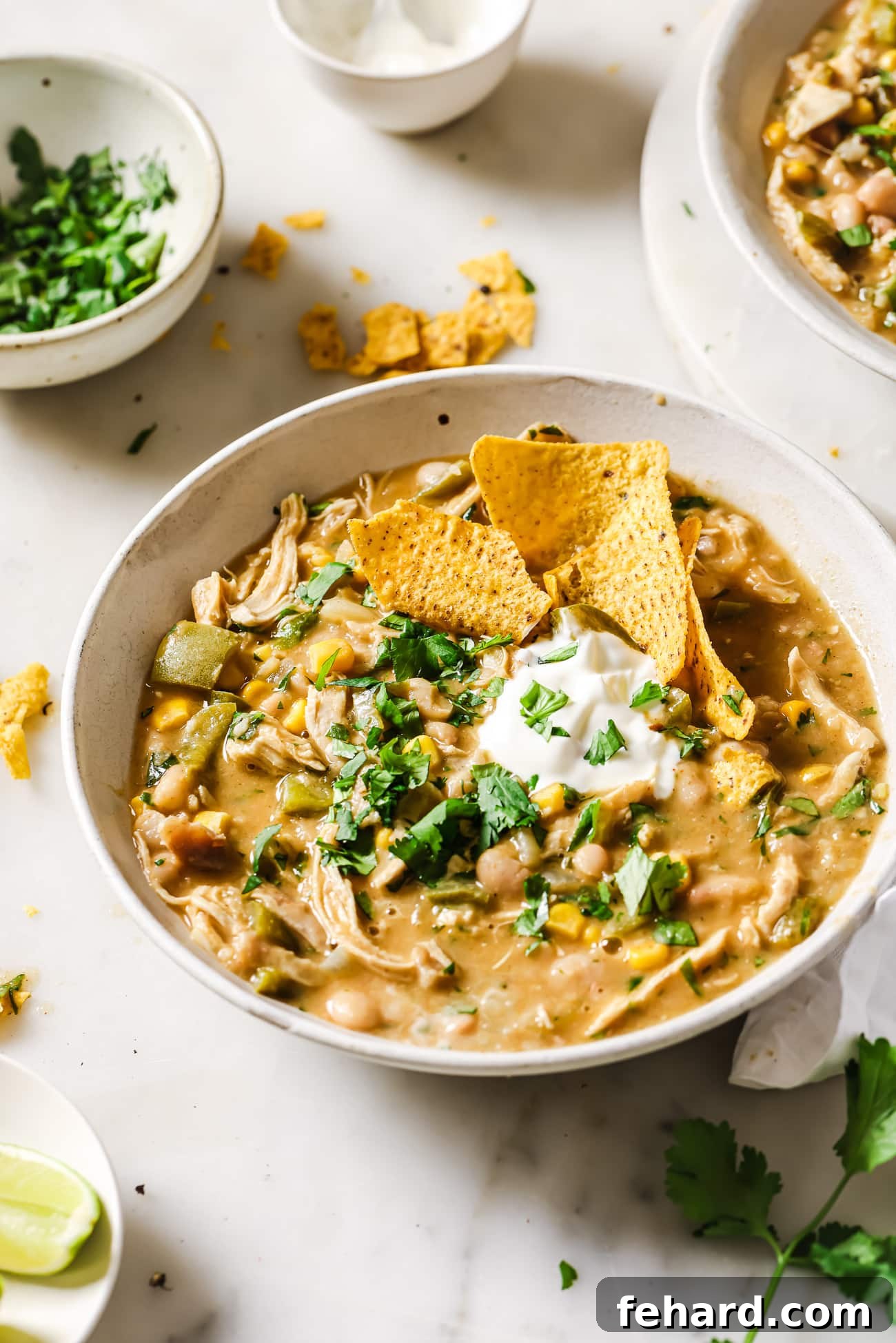A bowl of White Chicken Chili topped with sour cream, fresh cilantro, lime wedges, and a side of tortilla chips.