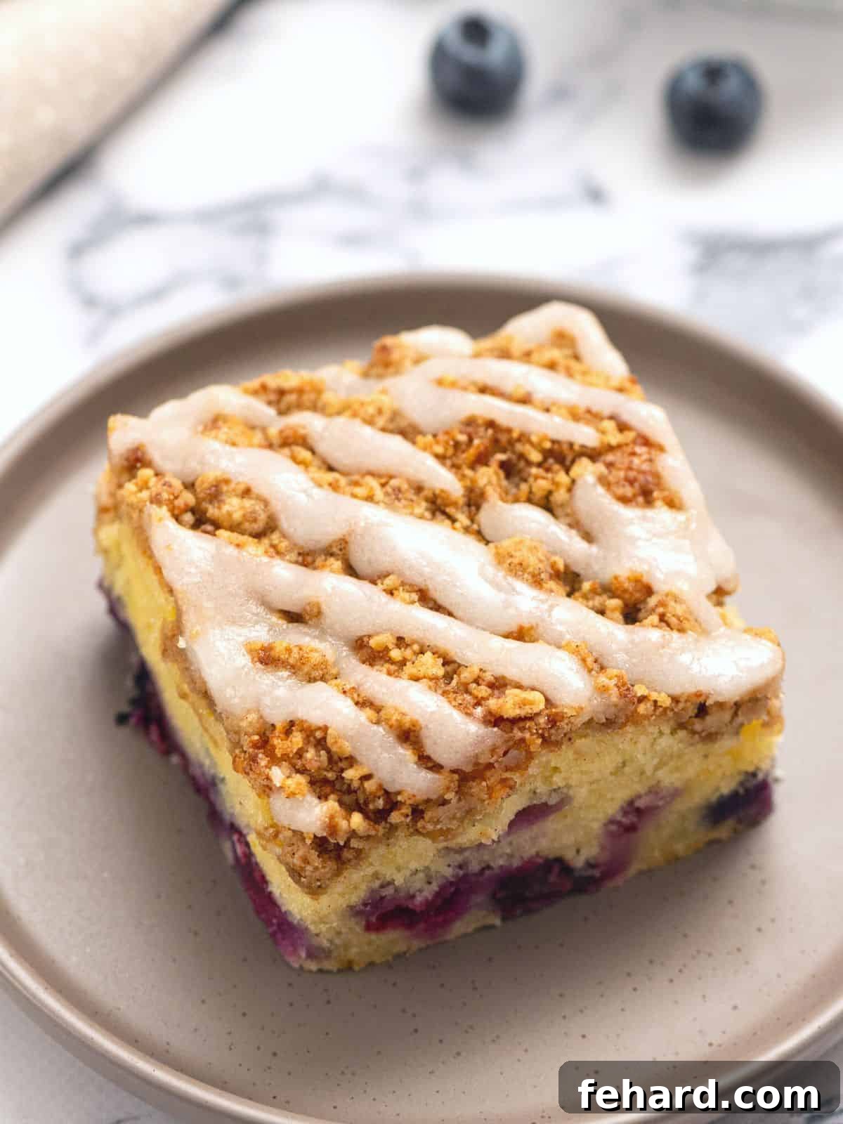 A perfectly sliced piece of Lemon Blueberry Coffee Cake, drizzled with a vibrant lemon glaze, resting on a white plate. Fresh blueberries and lemon slices are artfully arranged in the background, hinting at the cake's key ingredients.