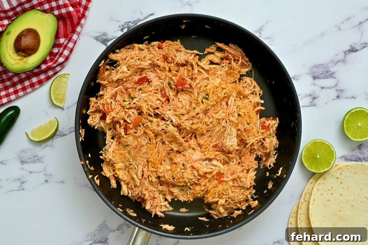 Close-up of shredded chicken simmering in a skillet with rich red salsa and a blend of aromatic taco seasonings.