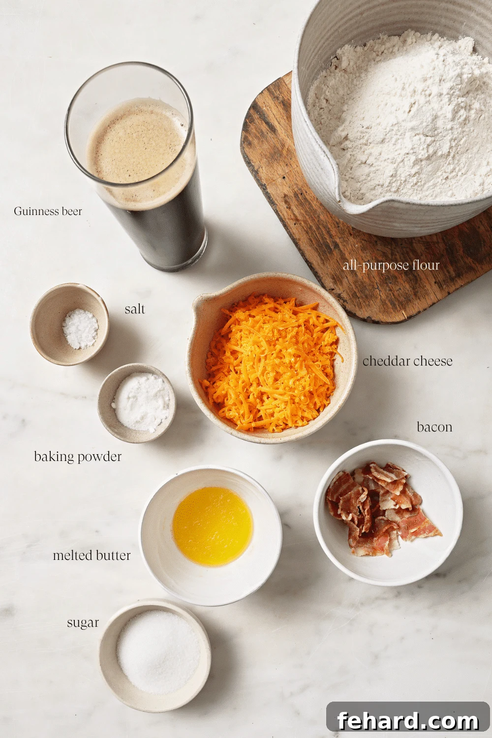 Pre-measured ingredients for homemade beer bread, including flour, baking powder, salt, sugar, shredded cheddar, Guinness beer, cooked bacon, and melted butter, all neatly arranged in small bowls.
