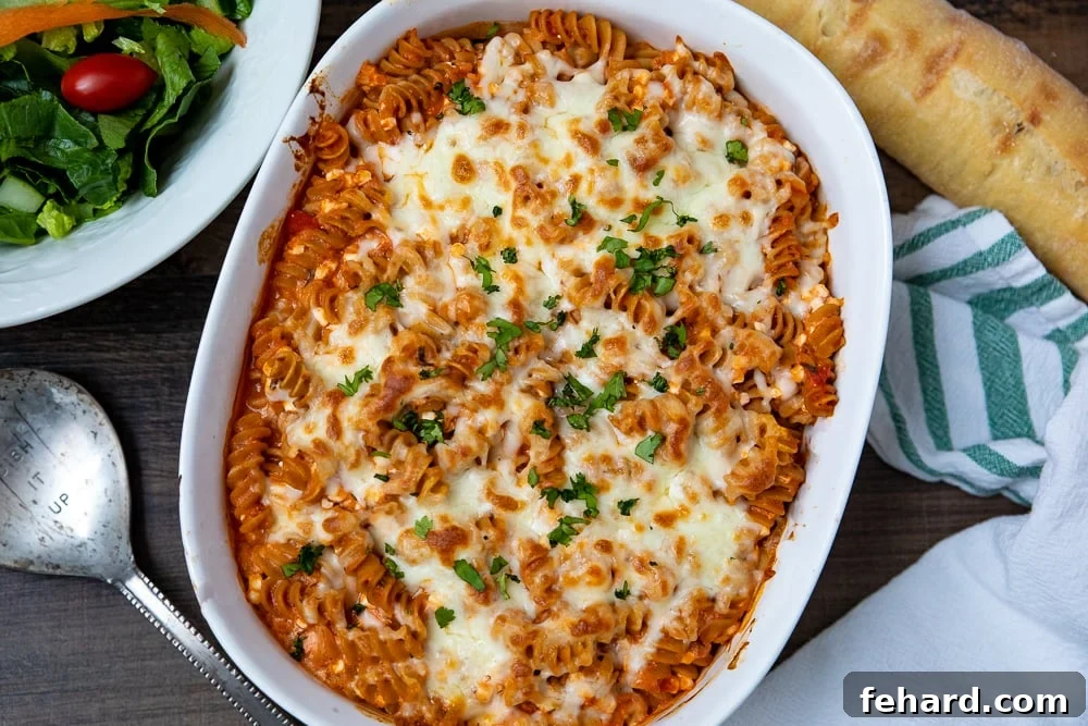 Close-up of a bubbling, cheesy Italian Baked Rotini casserole in a white dish, alongside a fresh tossed salad, ready to be served.