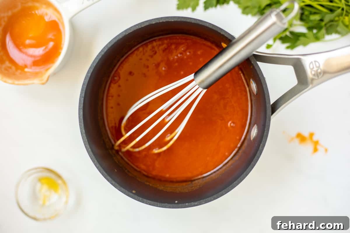 A white whisk is stirring honey into the newly made buffalo sauce in a saucepan, ensuring it's perfectly combined and smooth.