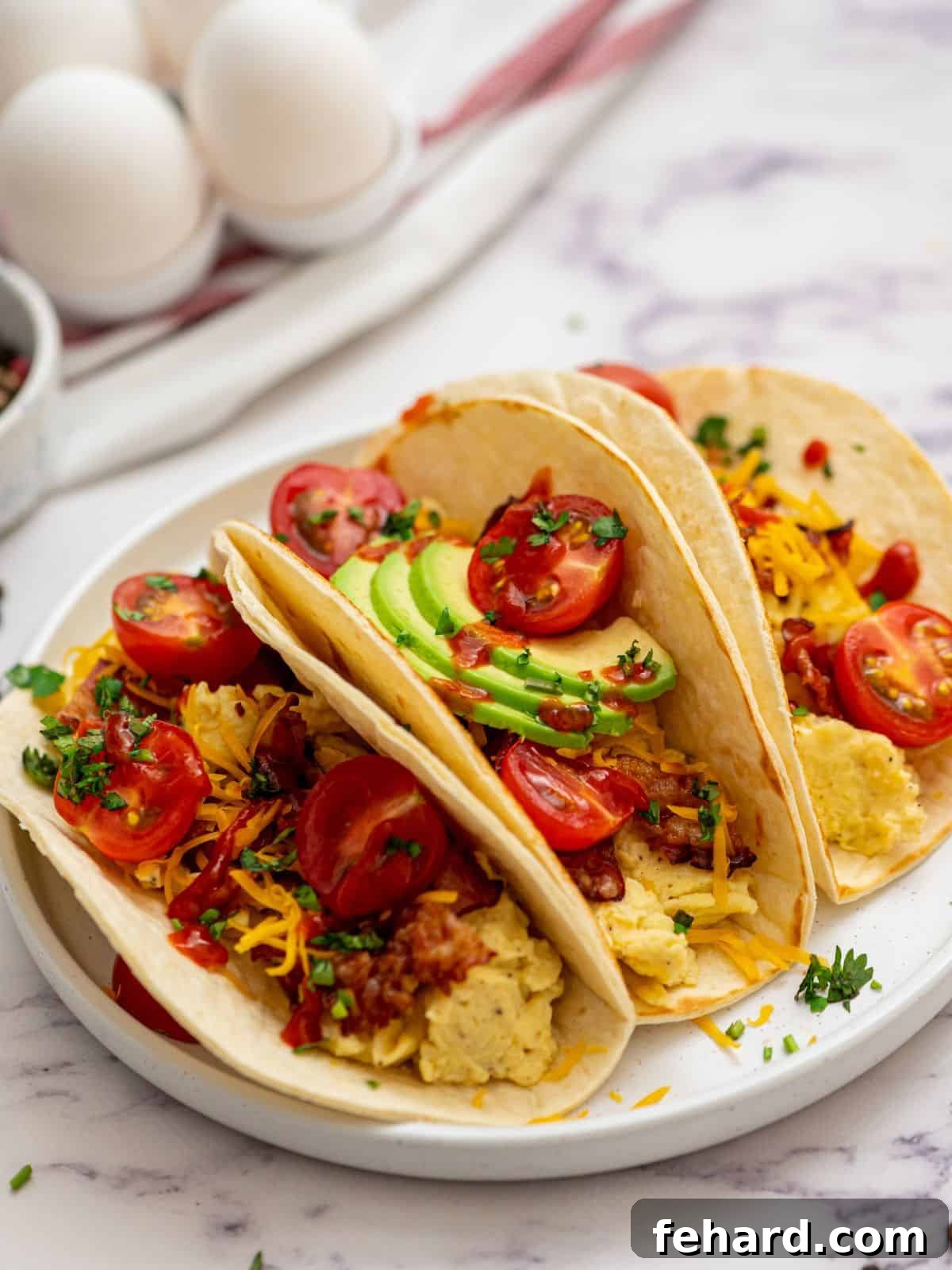 Three assembled breakfast tacos on a white plate, generously topped with scrambled eggs, crispy bacon, shredded cheese, fresh avocado slices, a drizzle of hot sauce, and diced tomatoes, ready to be enjoyed.