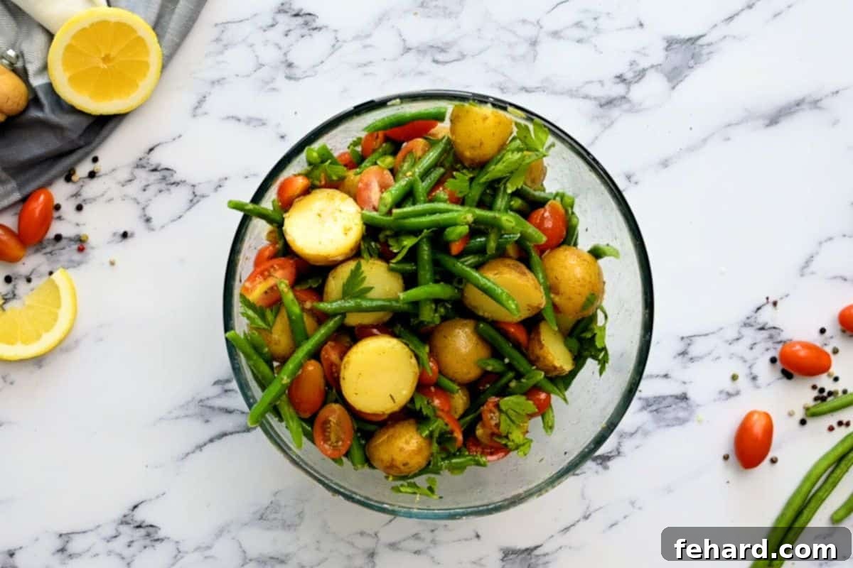All the vibrant ingredients for Italian Potato Salad, including potatoes, green beans, tomatoes, and parsley, gently tossed together in a large mixing bowl, coated in dressing.