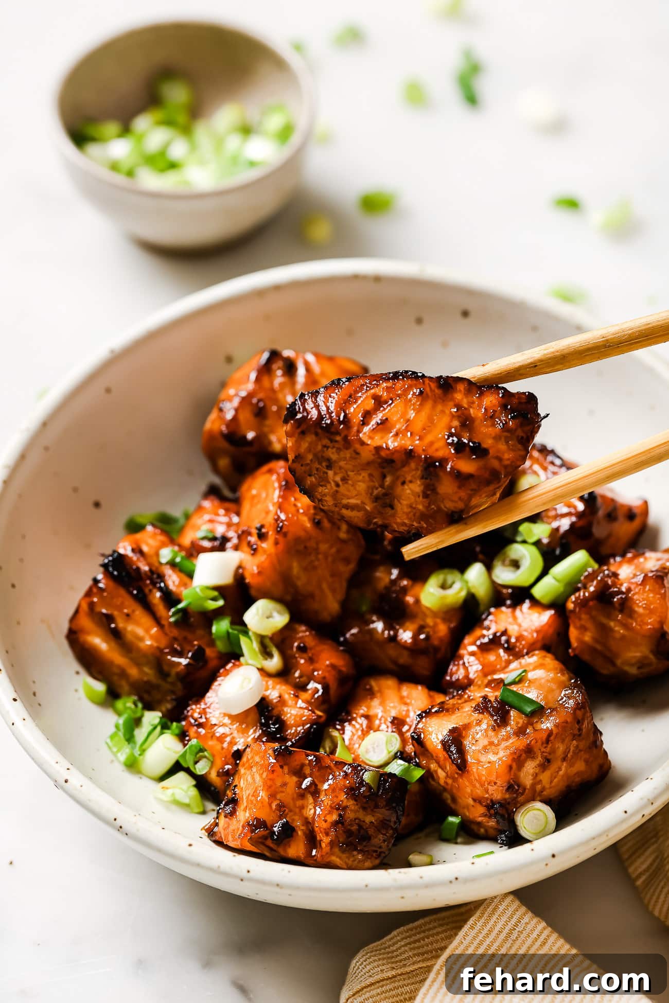 Chopsticks picking up a perfectly cooked air fryer salmon bite from a bowl, garnished with green onions and sesame seeds.