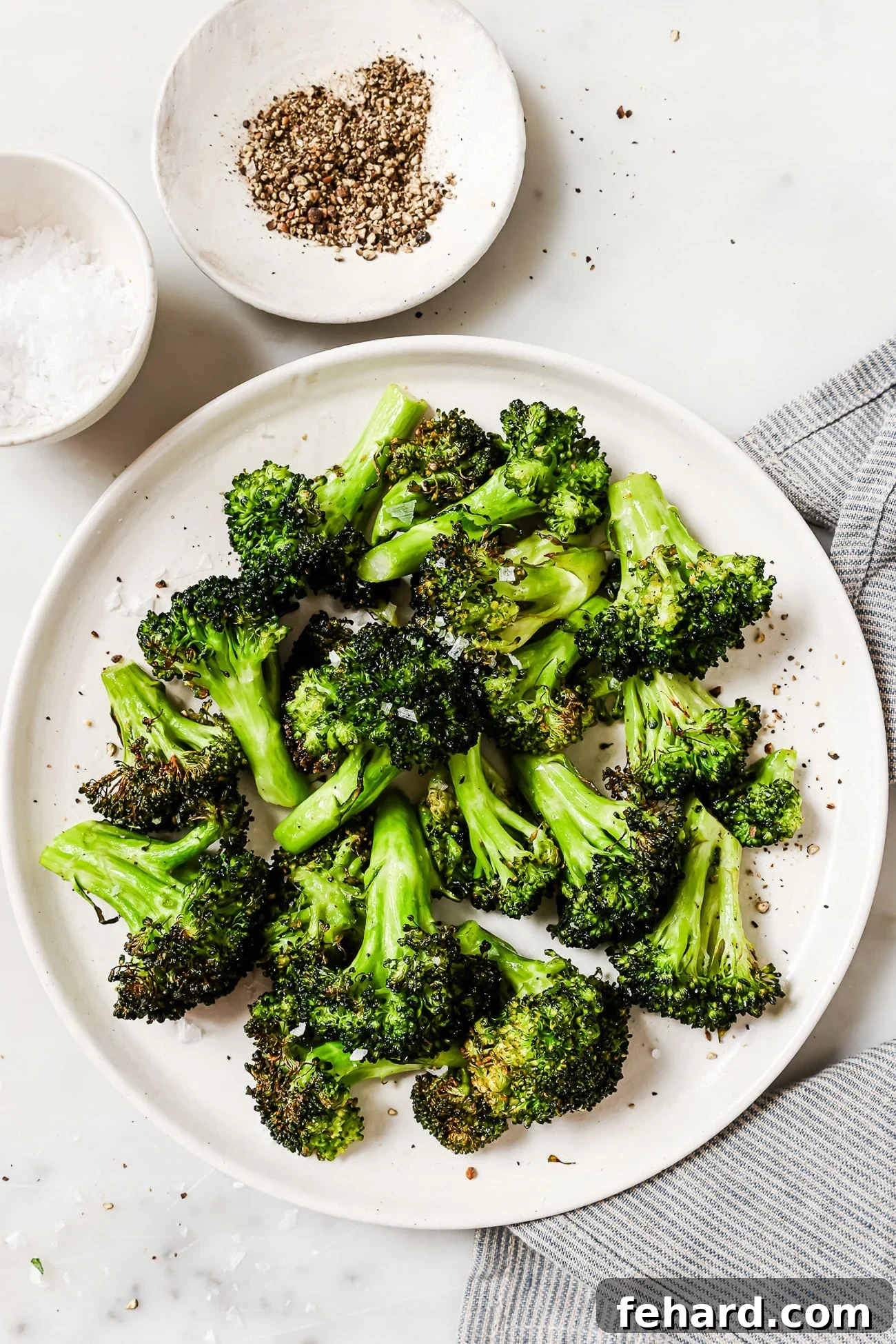 Quick & Crispy Air Fryer Broccoli 2 Air fryer broccoli on a white plate with salt and pepper in the background, showcasing its crispy texture.