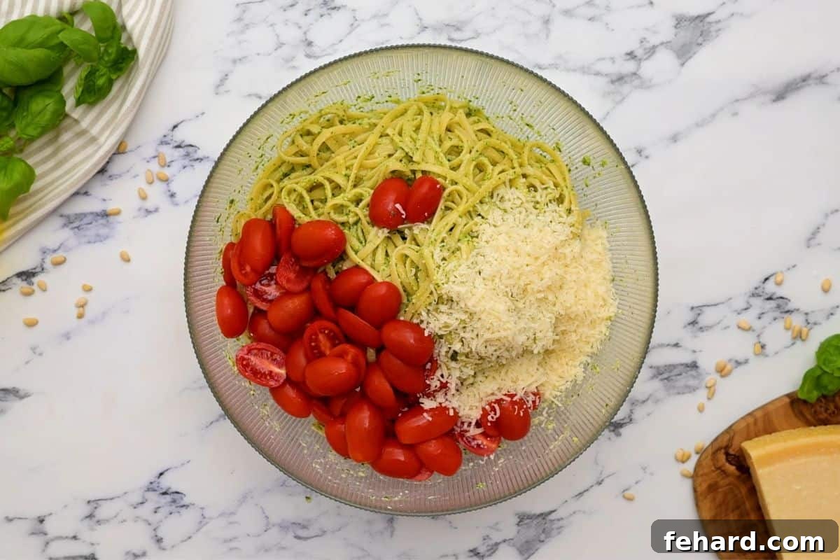 Large mixing bowl with cooked pasta, vibrant green pesto, red tomatoes, and shredded Parmesan cheese, ready to be served.