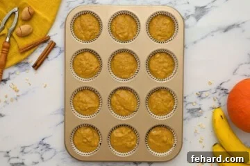 An ice cream scoop carefully measures and divides pumpkin muffin batter into a lined muffin tin.