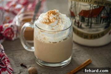 Pumpkin spice latte in clear glass much with maple syrup and cinnamon sticks in background.