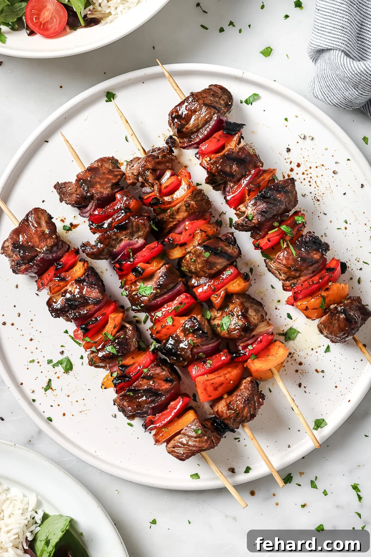 An overhead view of four beef kebabs on a white serving plate, garnished with fresh herbs.