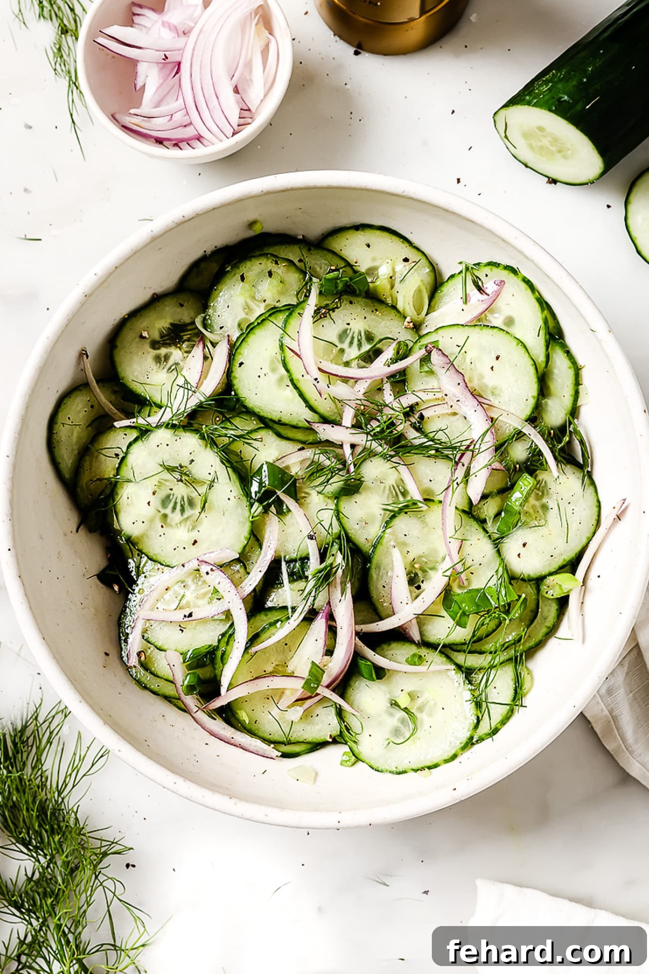 An overhead view of a large white serving bowl filled with fresh cucumber salad, garnished with dill.