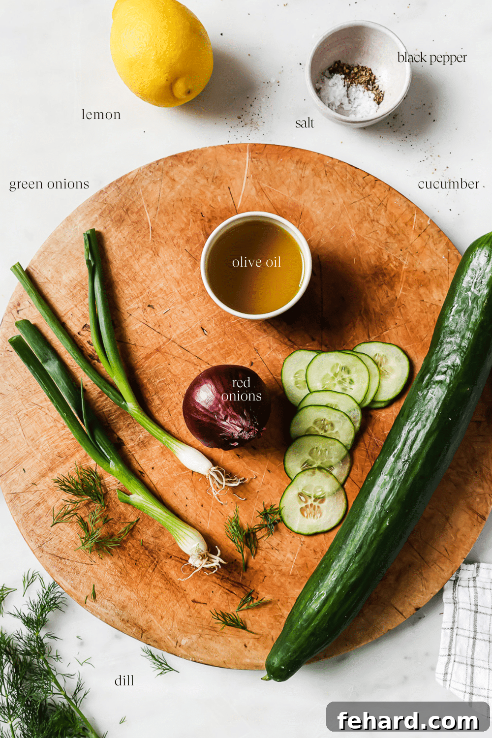 Fresh ingredients for cucumber salad, including sliced cucumbers, red onion, green onion, and a lemon, arranged on a white countertop and wooden cutting board.