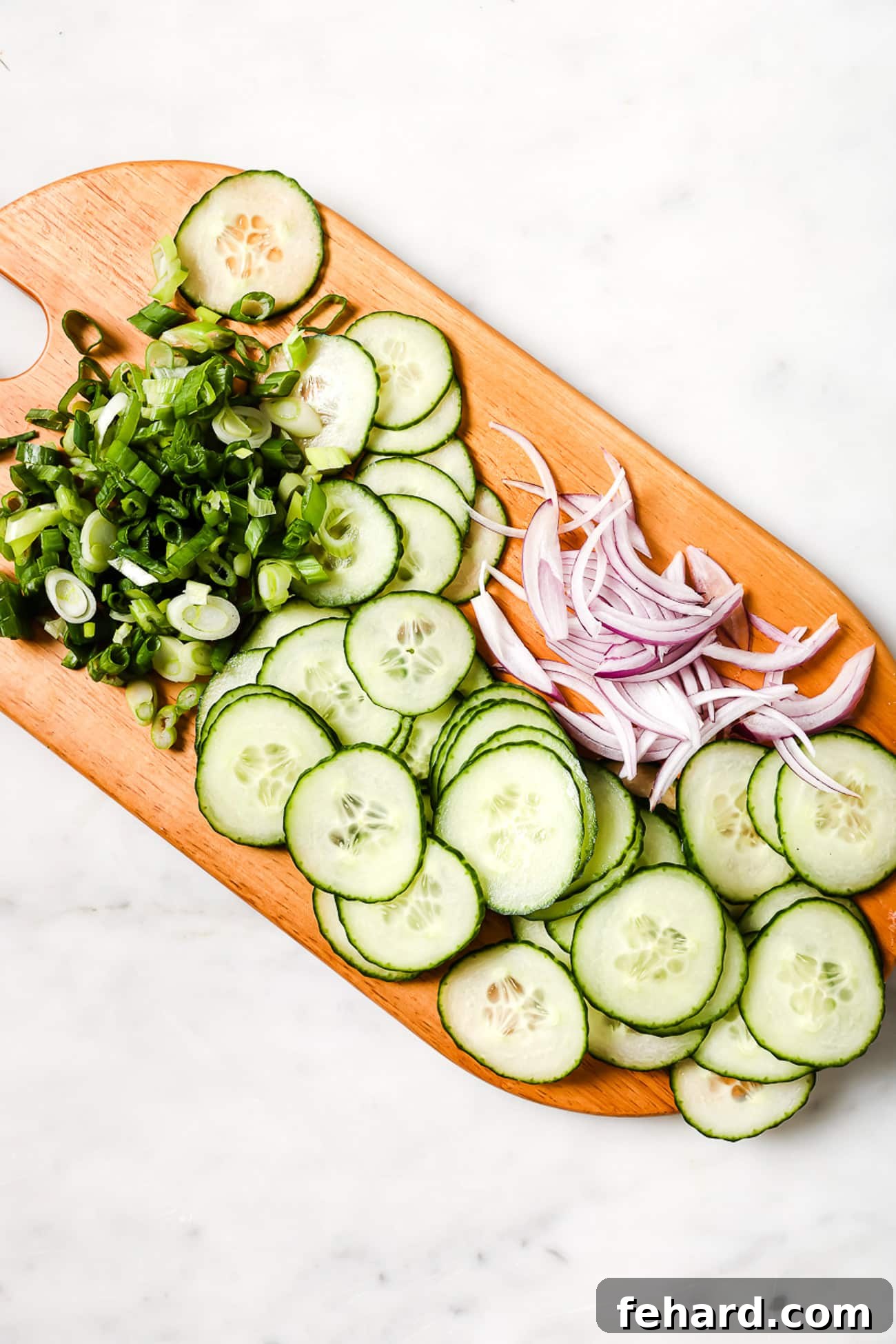 Thinly sliced cucumber salad ingredients, including cucumbers and red onion, neatly arranged on a wooden cutting board.