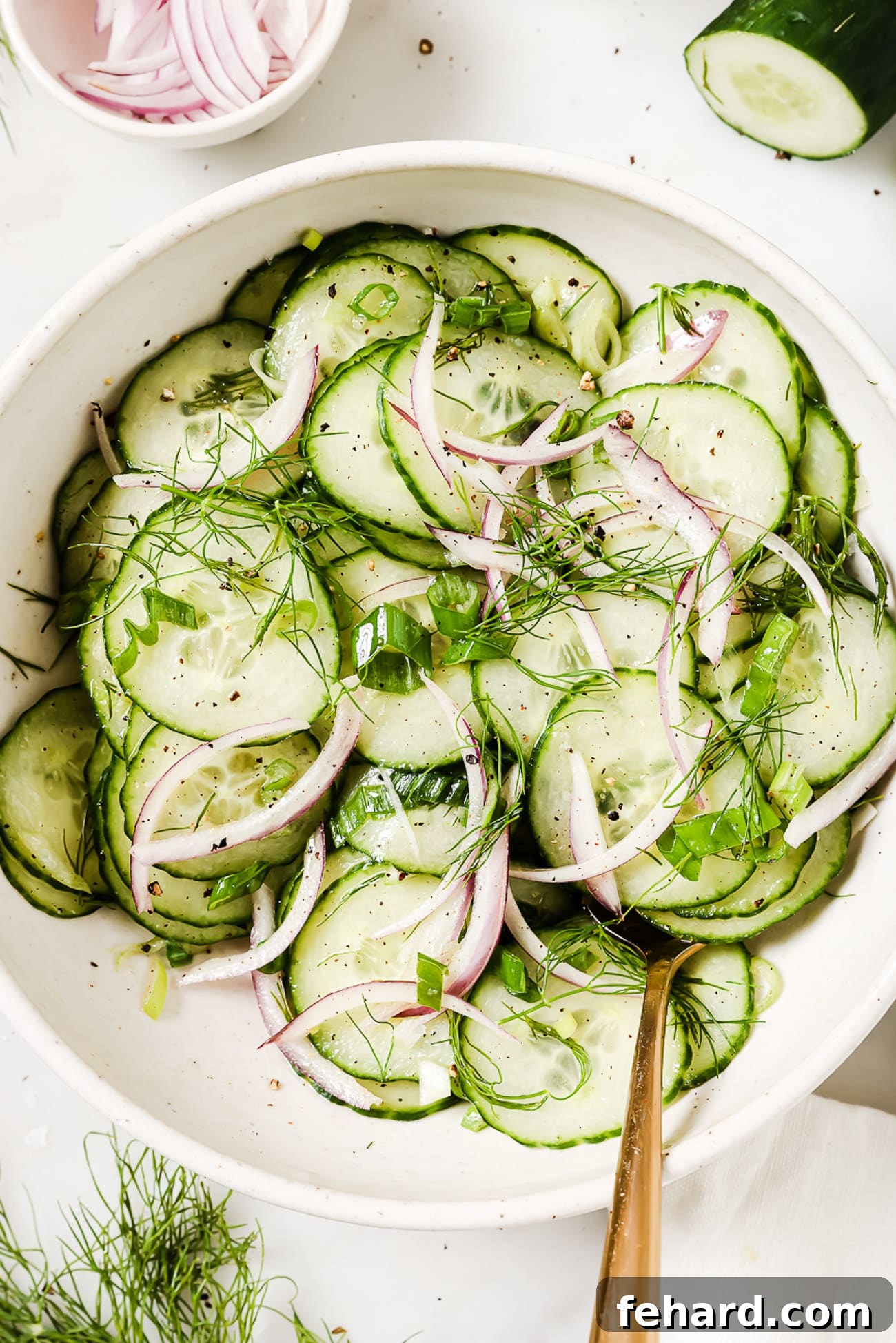 An overhead view of a fresh cucumber salad in a white bowl, accompanied by a gold spoon for serving.