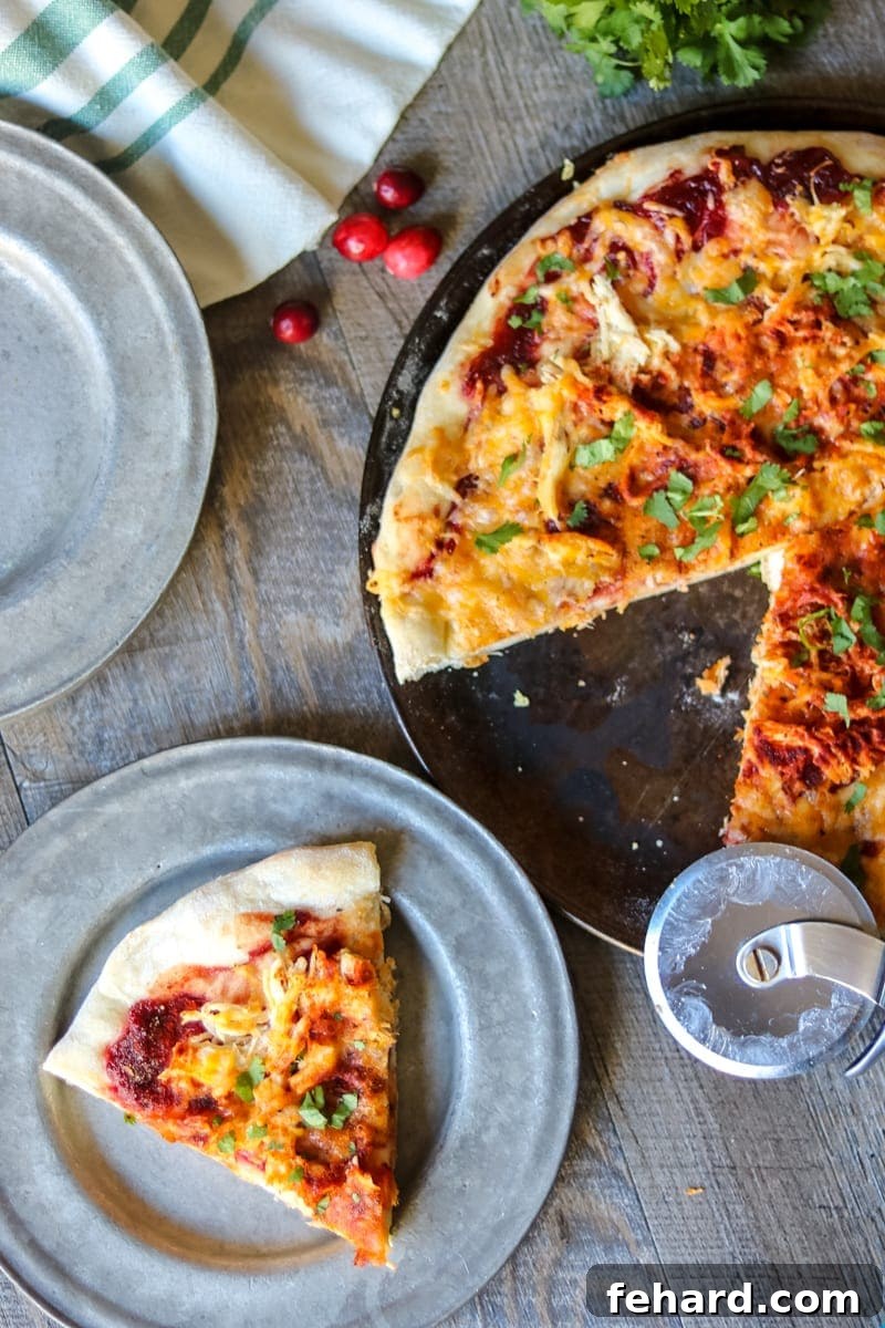 A delicious Thanksgiving Pizza on a pizza pan with one slice removed, showcasing melted cheese, turkey, and cranberry BBQ sauce. A single slice rests on a silver plate.