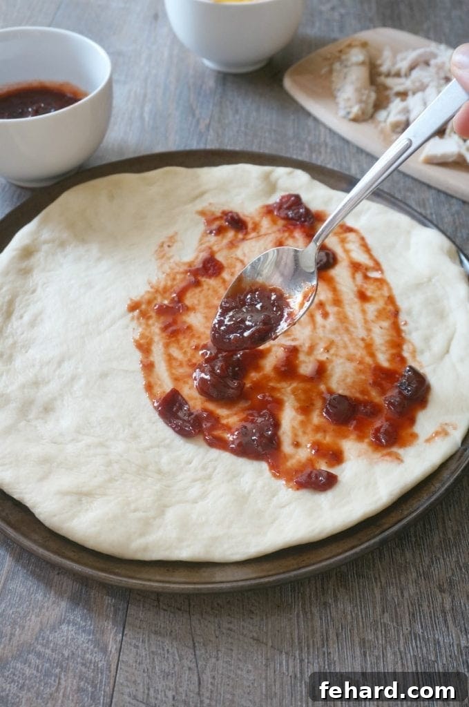 Applying cranberry BBQ sauce onto pizza dough with other toppings in the background, ready for assembly.