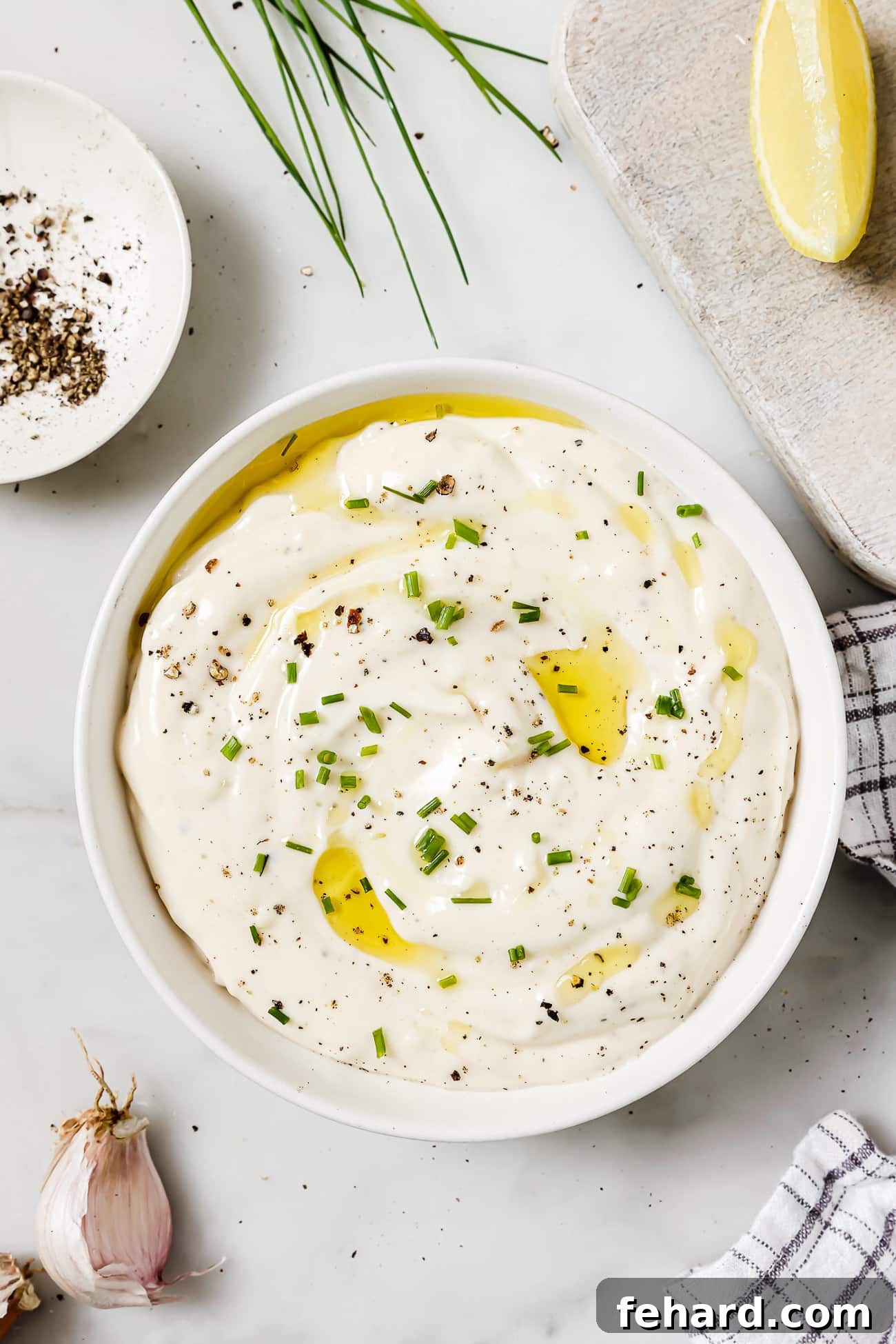 An overhead view of a rustic bowl of garlic aioli, artfully garnished with fresh herbs, with the individual ingredients subtly blurred in the background, creating an inviting culinary scene.