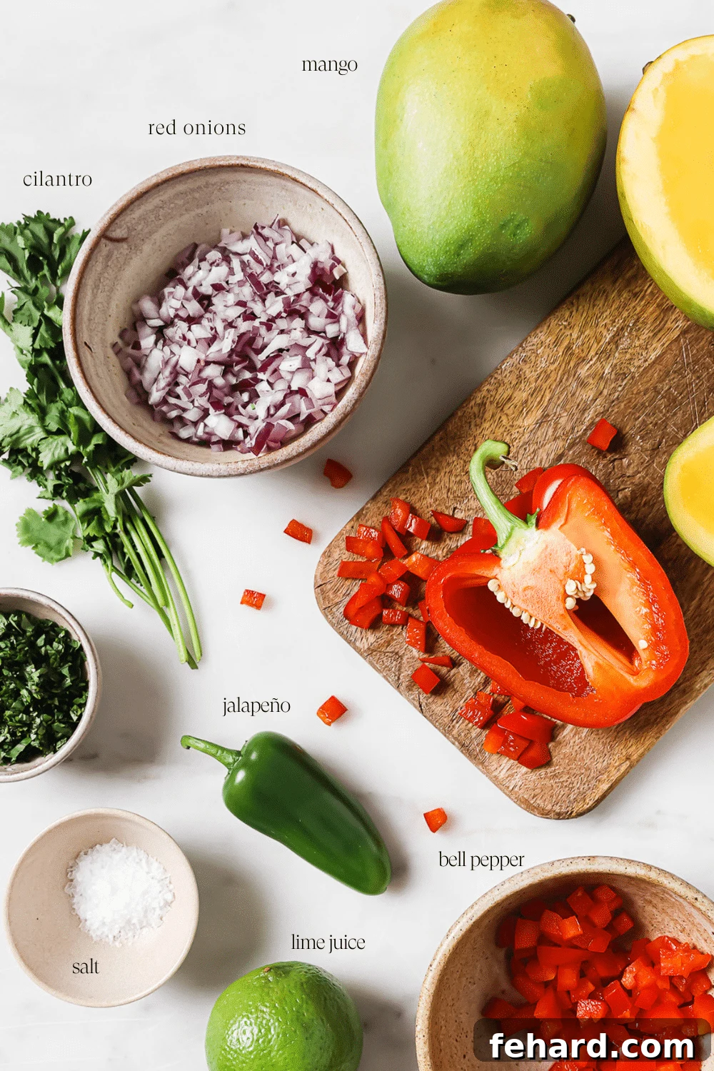 Sweet Heat Mango Salsa 3 An overhead view of the fresh ingredients for mango salsa, including diced mangoes, red bell pepper, red onion, cilantro, and jalapeño, neatly displayed on a wooden cutting board and white countertop, ready for preparation.