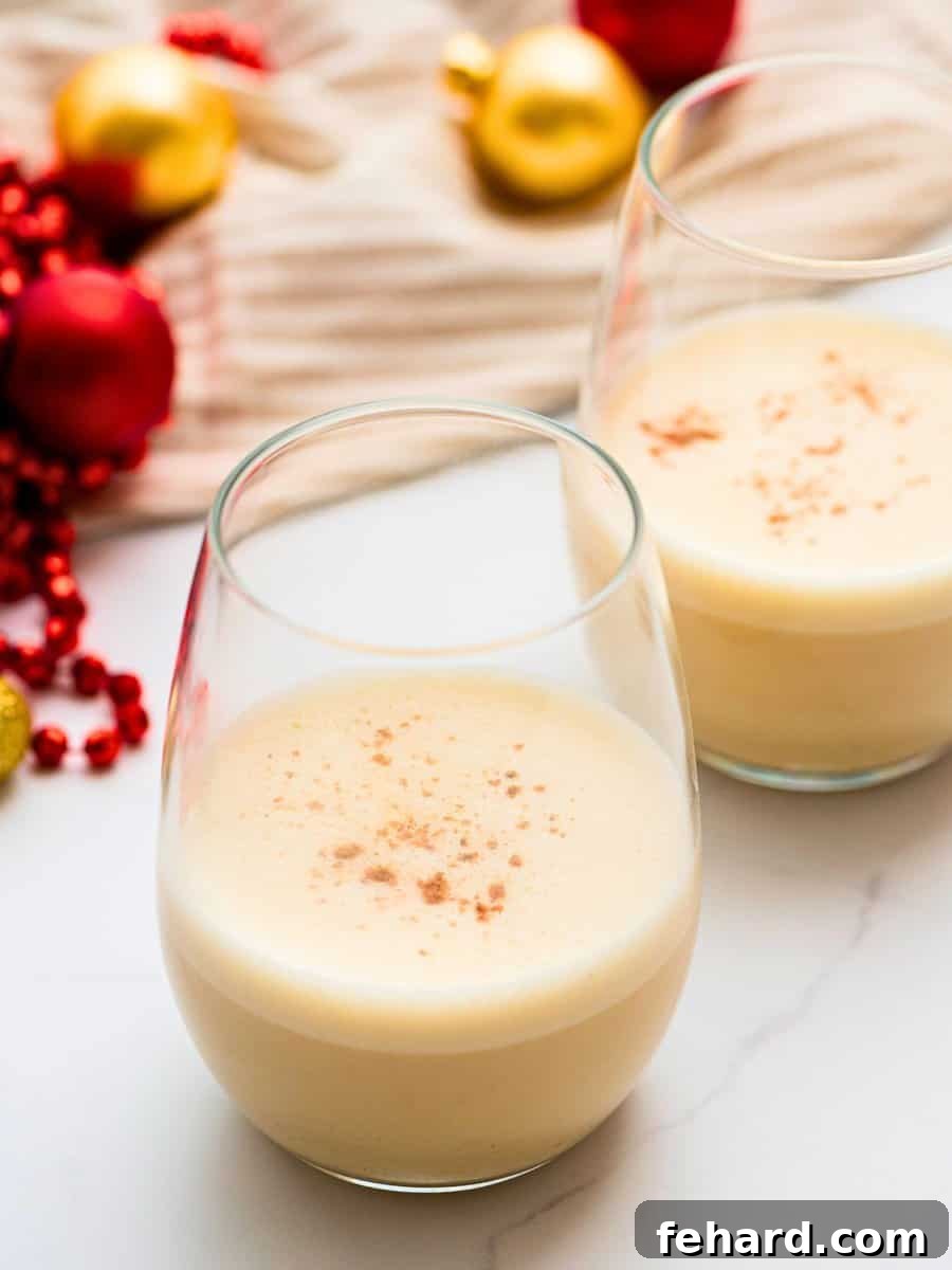 Two glasses of creamy dairy-free eggnog, made with coconut milk and topped with grated nutmeg, next to festive Christmas ornaments.