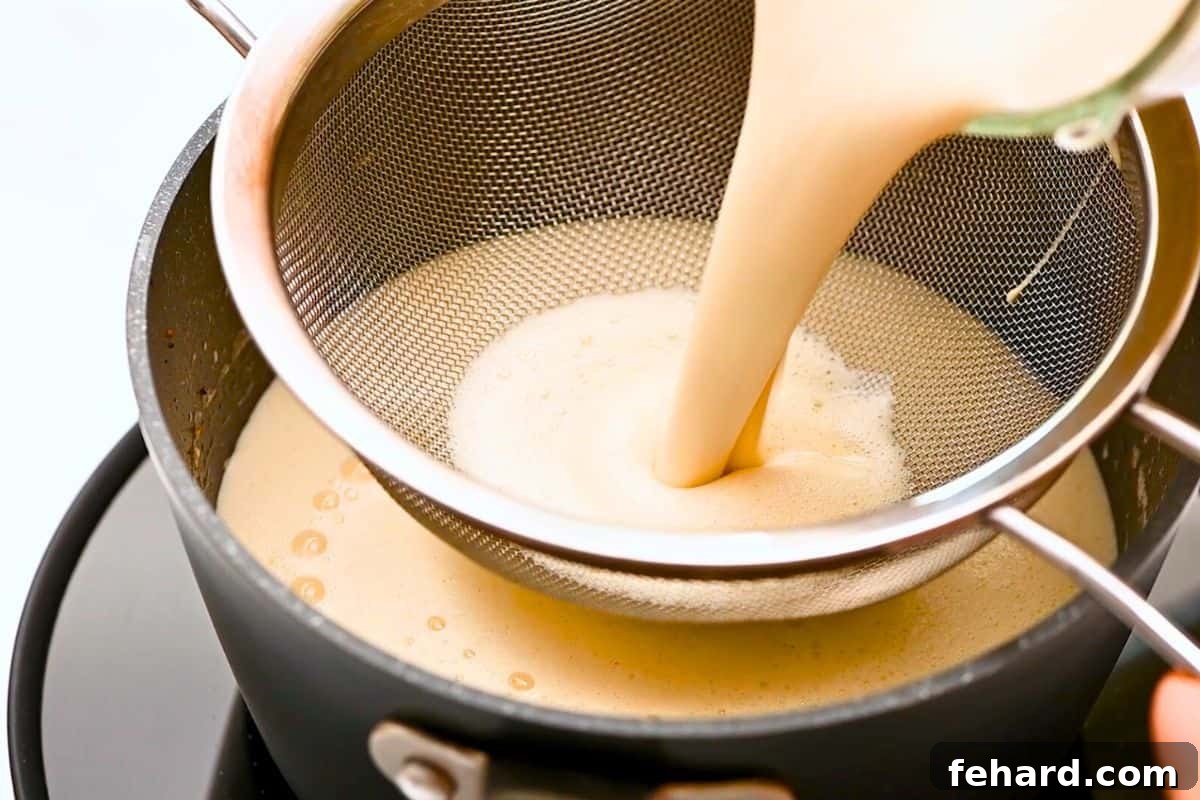 Blended eggnog being poured through a fine mesh strainer into a saucepan to remove any potential lumps.