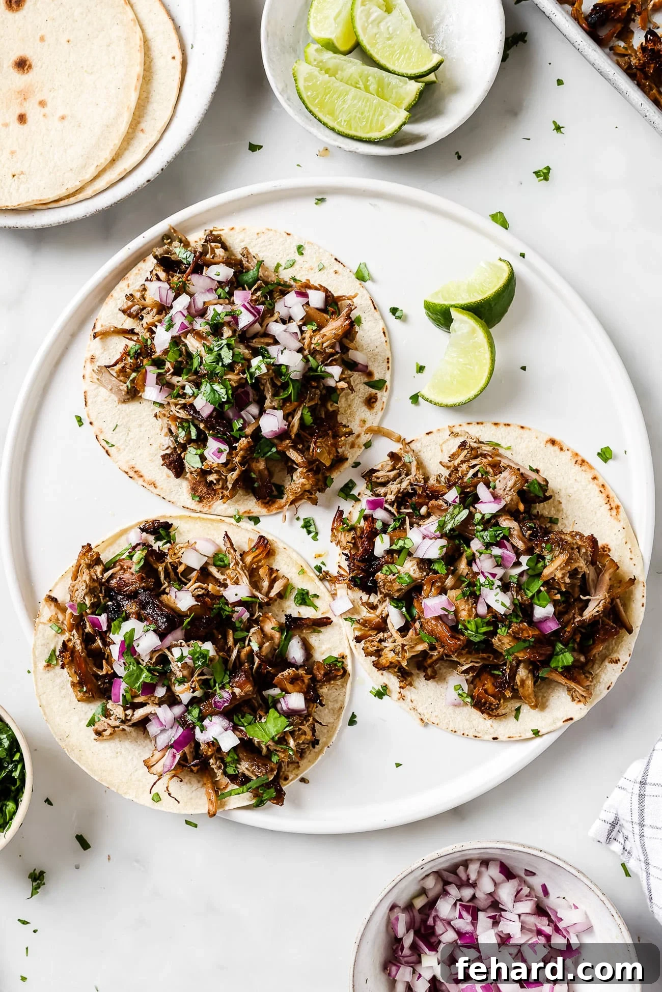 An overhead view of three perfectly assembled carnitas tacos, garnished with red onion and cilantro, served on a white plate with fresh lime wedges on the side.