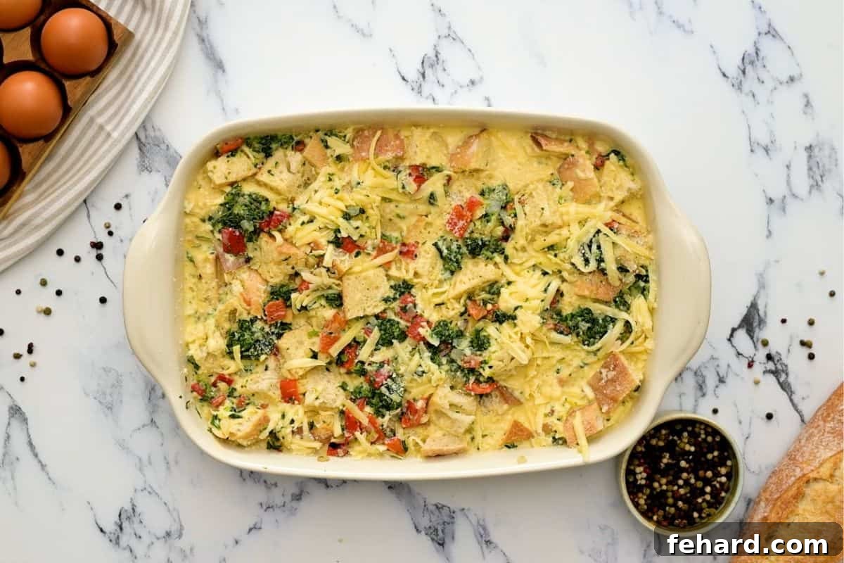 Bread, eggs, spinach, and roasted red peppers in baking dish before baking breakfast strata.