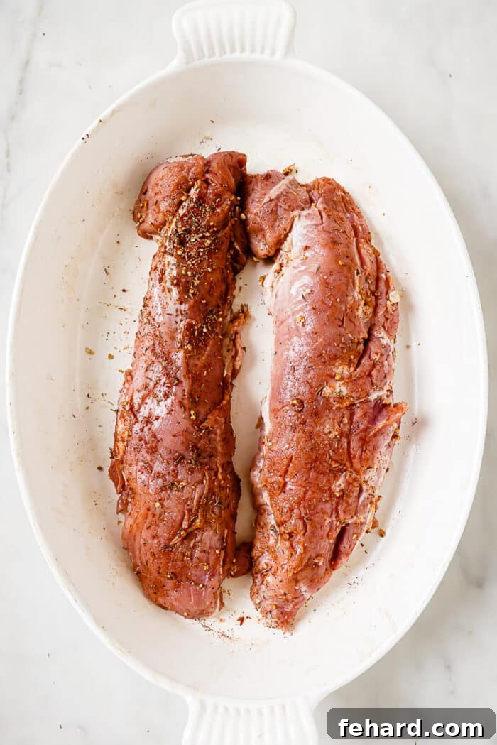 Two seasoned pork tenderloins resting in a sturdy baking dish, coated evenly with a flavorful dry rub, awaiting the next cooking stage.