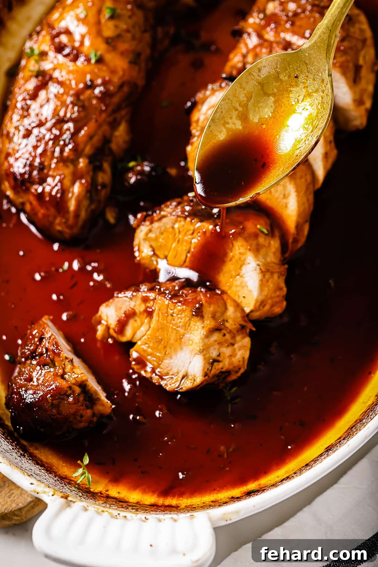A hand holding a spoon artfully drizzles rich, shimmering honey garlic sauce over slices of perfectly cooked pork tenderloin, emphasizing its tender texture and delectable glaze.