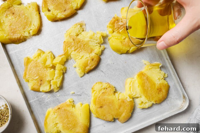 Crispy Smashed Potatoes 6 A hand drizzling golden olive oil generously over the smashed potatoes spread out on a baking sheet.