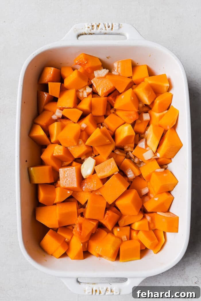 Raw, perfectly diced butternut squash, along with chopped shallots and garlic, waiting in a white casserole dish before roasting.