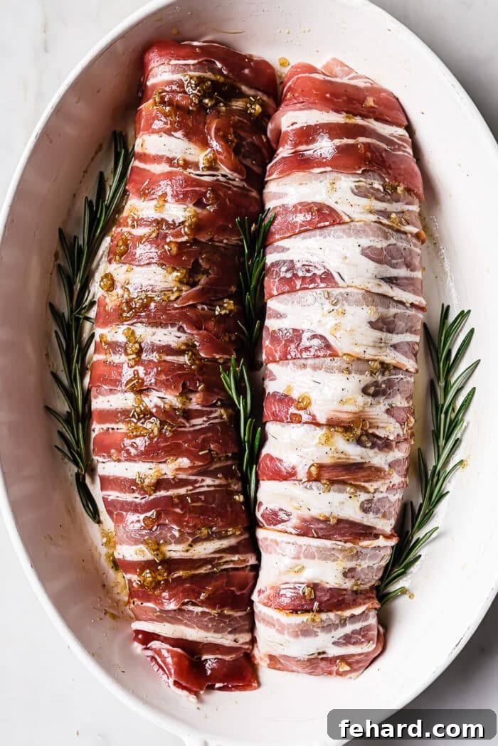 Two bacon wrapped pork tenderloins in a white baking dish with rosemary sprigs around it. 