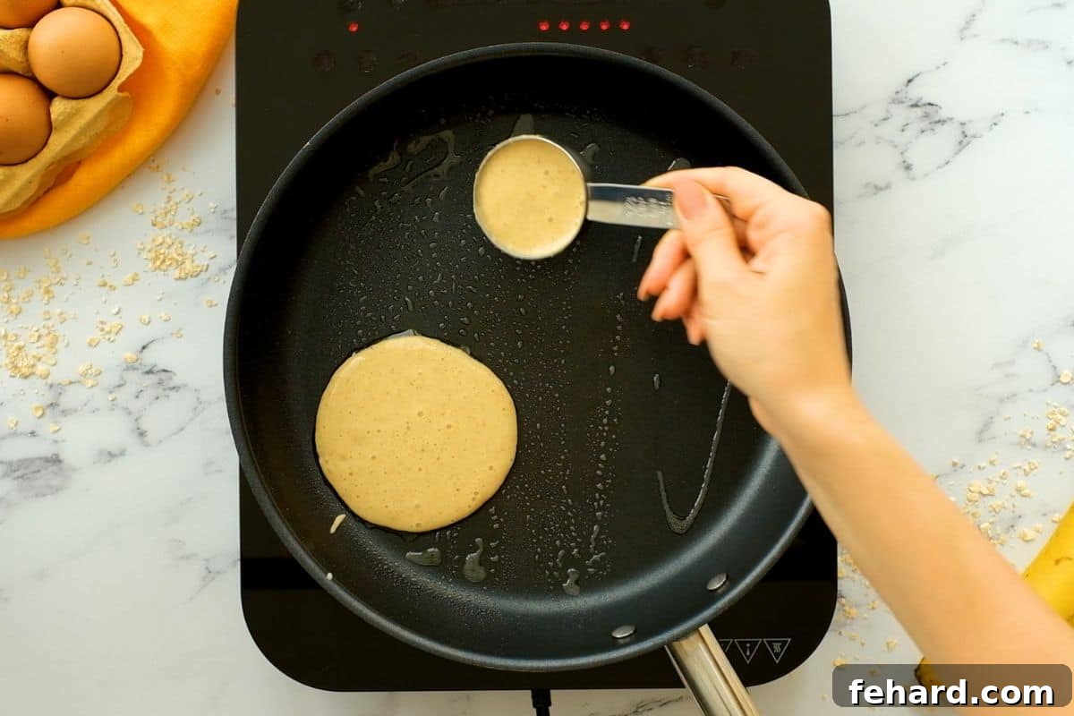 A quarter-cup measuring cup expertly scooping oatmeal banana pancake batter onto a hot, greased skillet, ensuring uniform pancake size.
