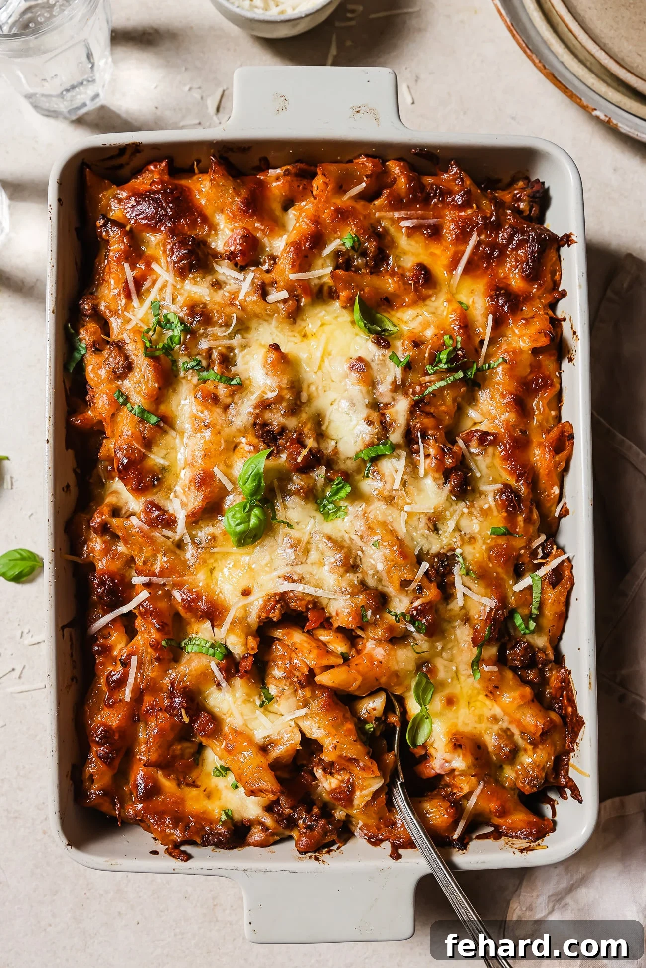 Baked ziti with ground beef in a white casserole dish garnished with basil leaves and Parmesan cheese. 
