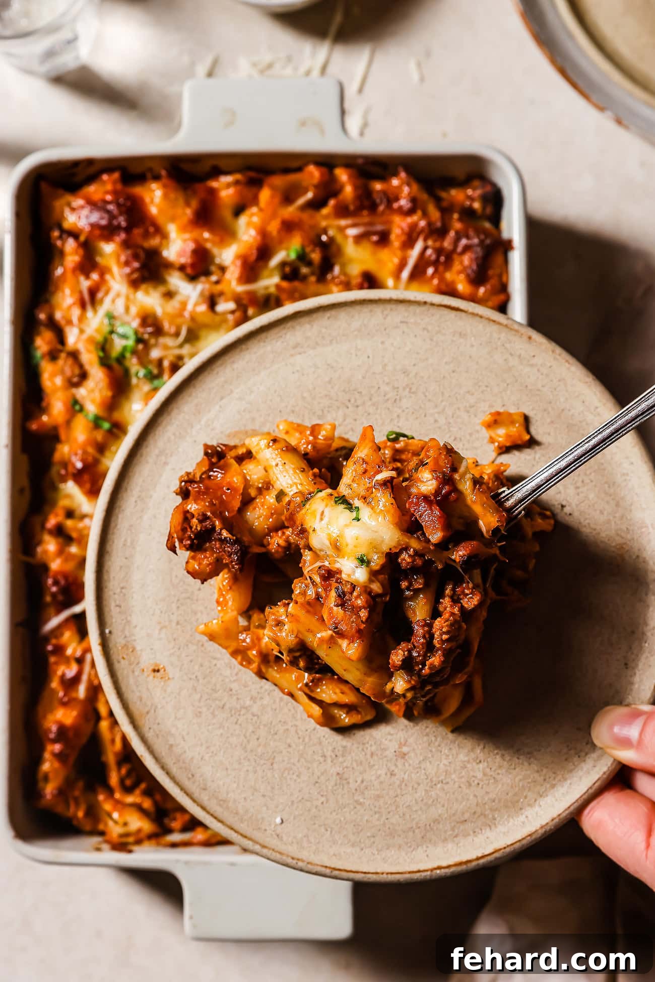 A serving of baked ziti with ground beef on a plate with the baking dish in the background. 