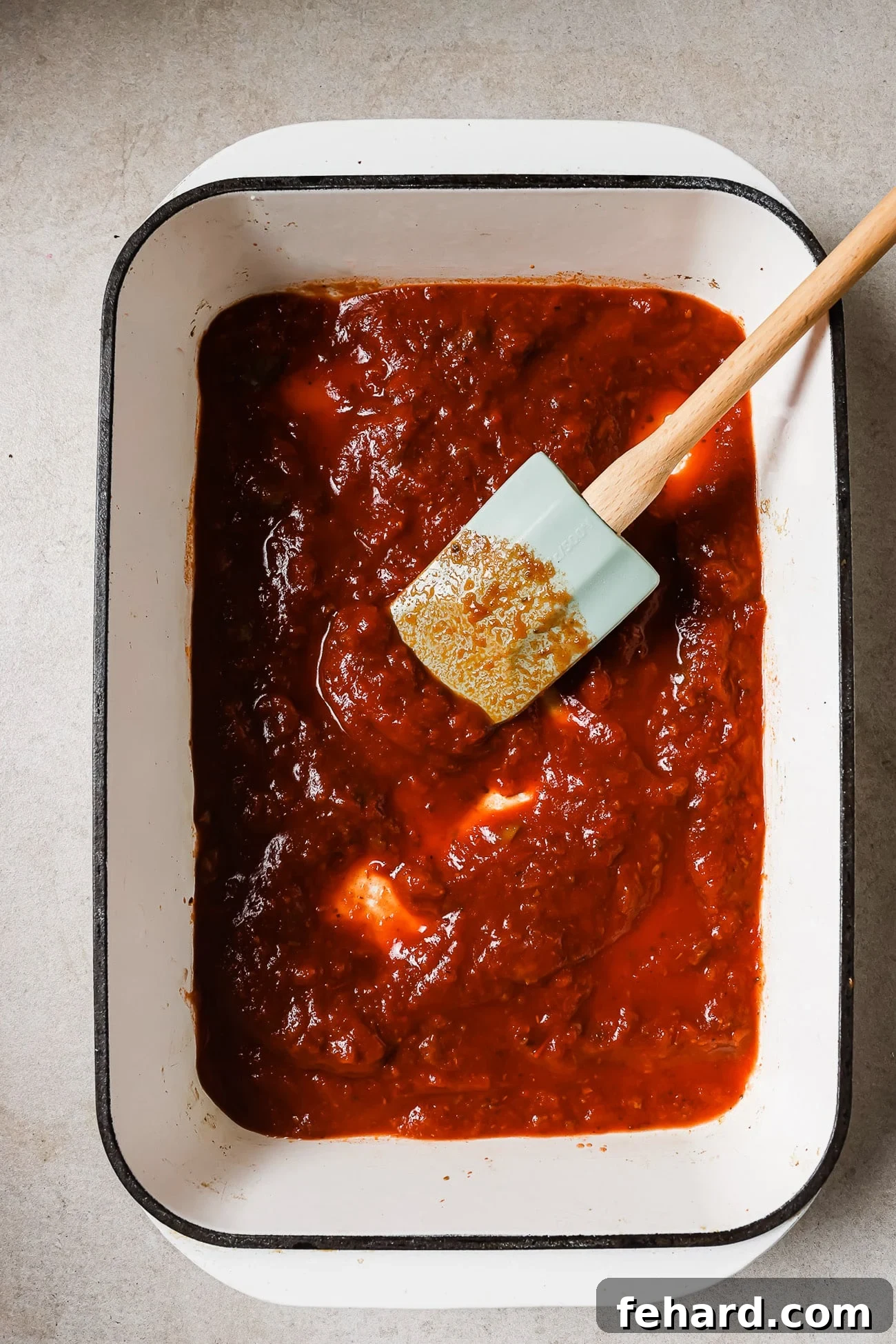 A white casserole dish with marinara sauce and a blue spatula in it. 