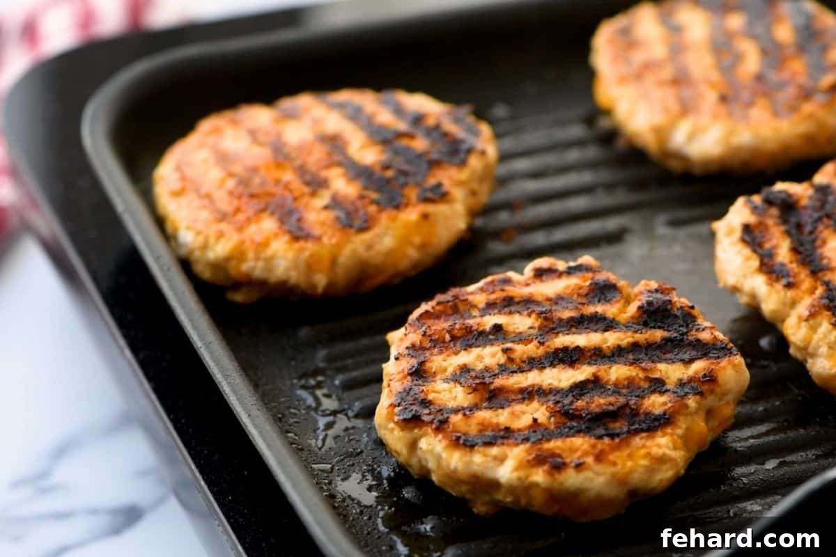 Four chicken burgers sizzling on a hot grill, showing a golden-brown crust forming.