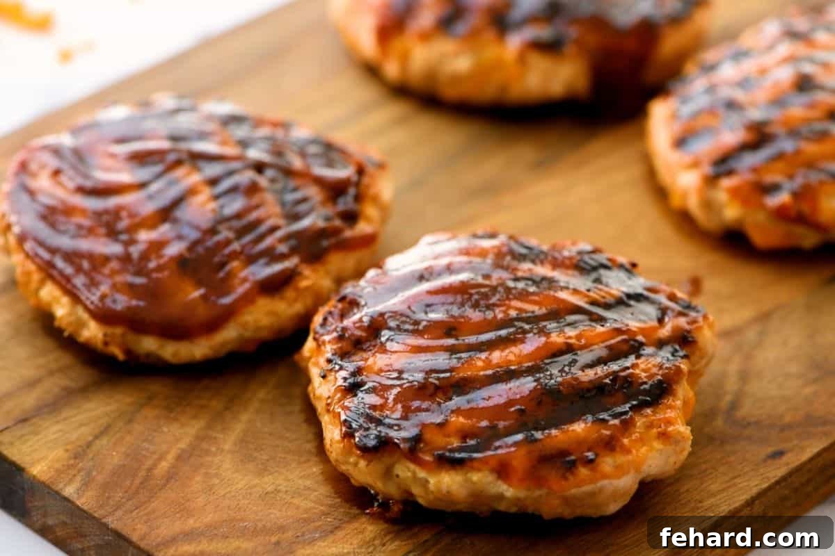 Four finished BBQ chicken burgers, glazed with barbecue sauce, resting on a wooden platter.
