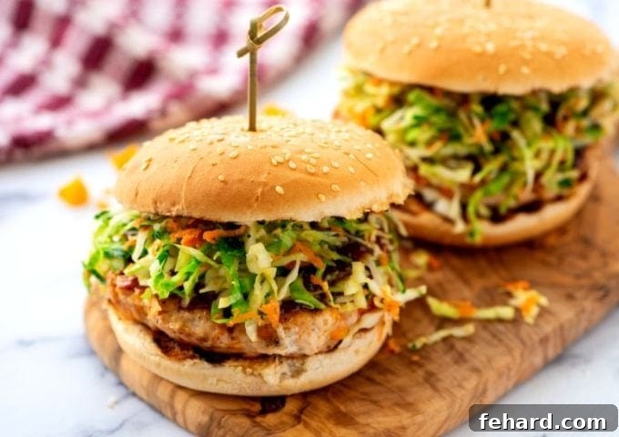 A fully assembled BBQ chicken burger on a bun with coleslaw, ready to eat, alongside another burger on a cutting board.