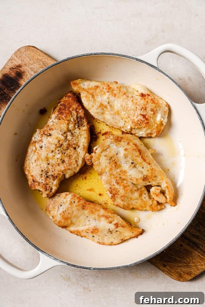 Sticky Honey Garlic Chicken Breast 5 An overhead view of four golden-brown seared chicken cutlets cooking in a white skillet over medium-high heat.