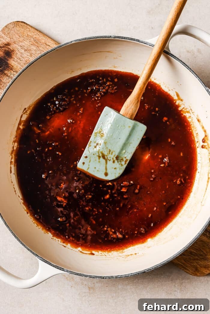 Sticky Honey Garlic Chicken Breast 7 Sticky honey garlic sauce simmering in a white skillet, being stirred with a blue rubber spatula, showing its thick and glossy texture.