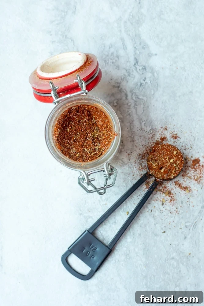 A glass spice jar filled with freshly made homemade Cajun seasoning, with a small measuring spoon beside it, suggesting readiness for use.