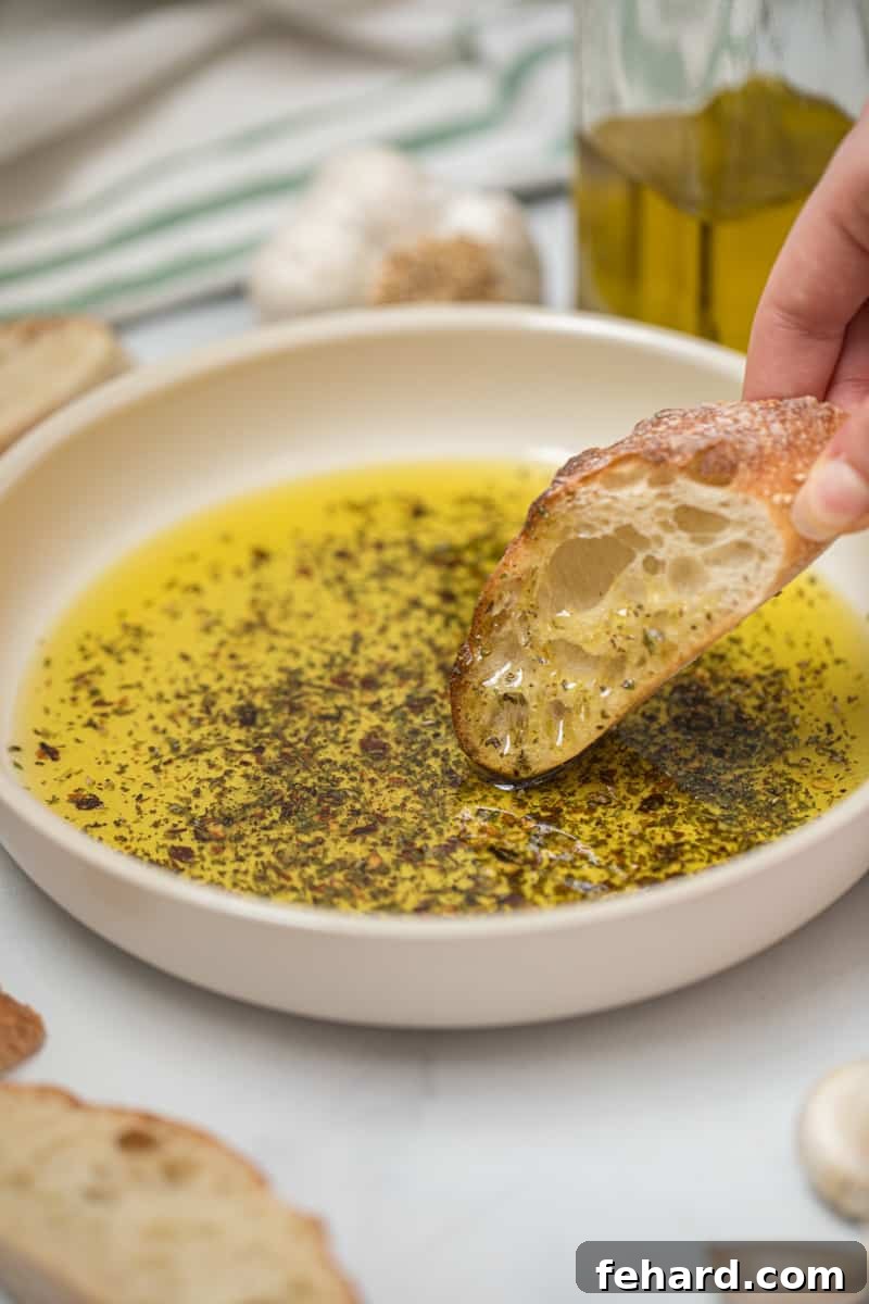 Toasted baguette being dipped into olive oil bread dip with fresh garlic in background.