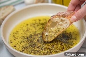 Toasted baguette being dipped into olive oil bread dip with fresh garlic in background.